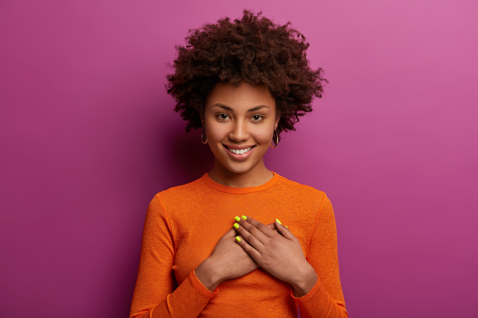 Pleased girl in orange jumper presses palms to heart, makes thankful gesture, touched with cordial congratulations, smiles positively, isolated over purple background. Acknowledgement concept